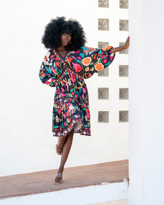 Woman in a colorful dress standing on a wooden deck with a white wall and geometric patterned wall in the background.