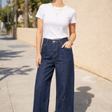 Woman wearing a white t-shirt and blue jeans standing on a sidewalk with palm trees in the background.