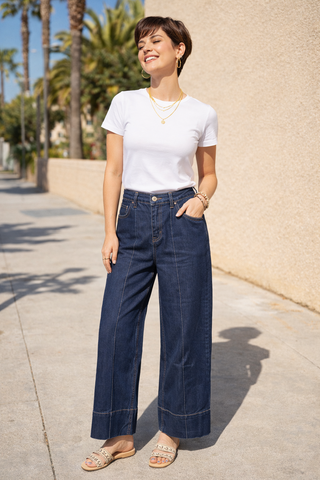 Woman wearing a white t-shirt and blue jeans standing on a sidewalk with palm trees in the background.