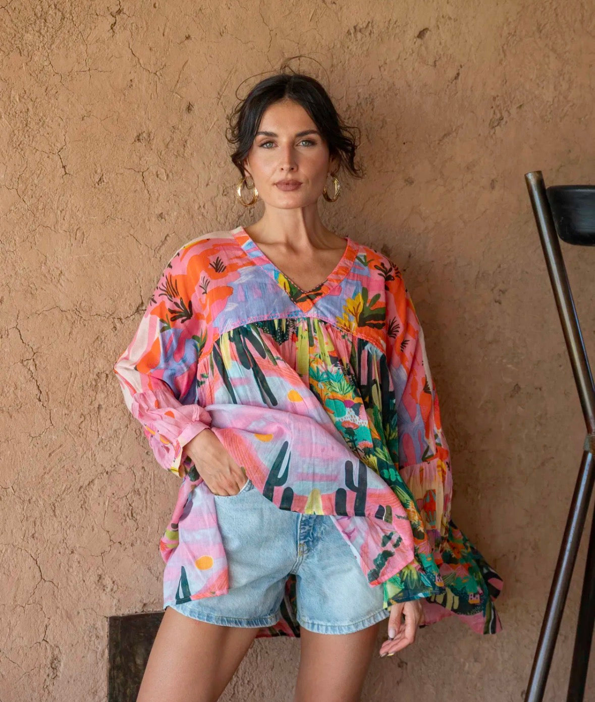 Woman wearing a colorful patterned top and denim shorts against a beige wall.
