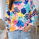 Person wearing a colorful floral and leopard print top with denim shorts indoors.
