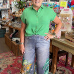 Woman in a green shirt and floral pants standing in a store with various items on shelves.