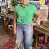 Woman in a green shirt and floral pants standing in a store with various items on shelves.