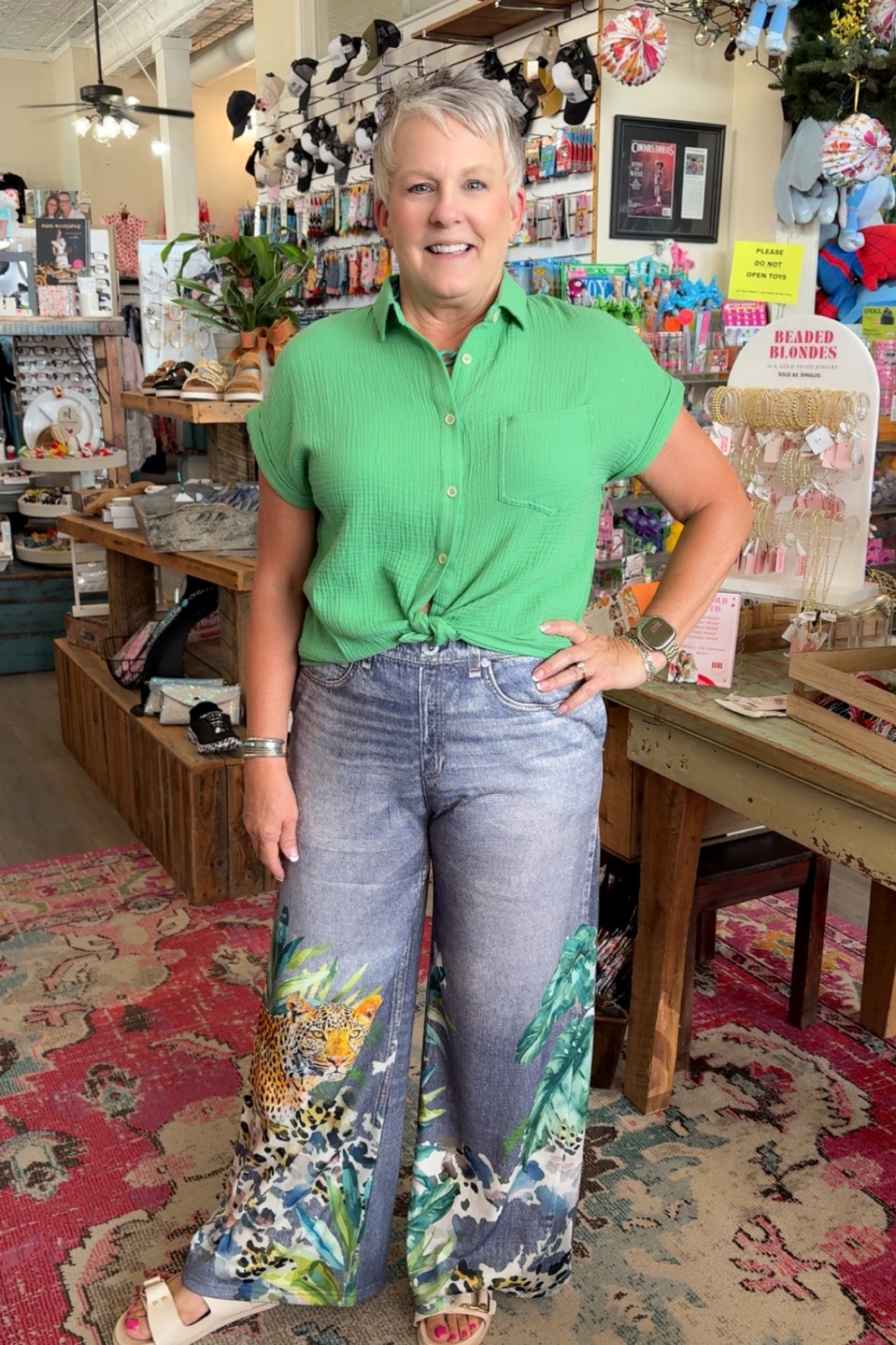 Woman in a green shirt and floral pants standing in a store with various items on shelves.