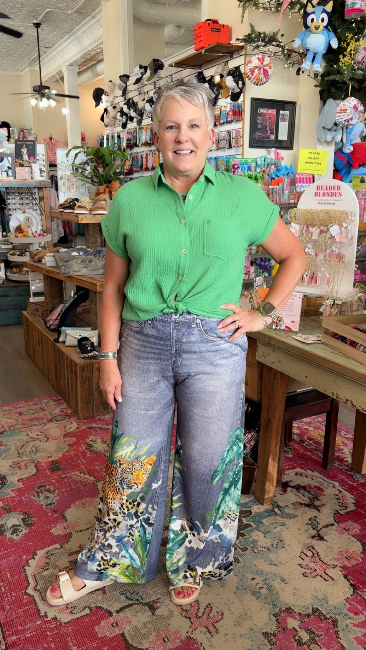 Woman in a green shirt and floral pants standing in a store with various items on shelves.