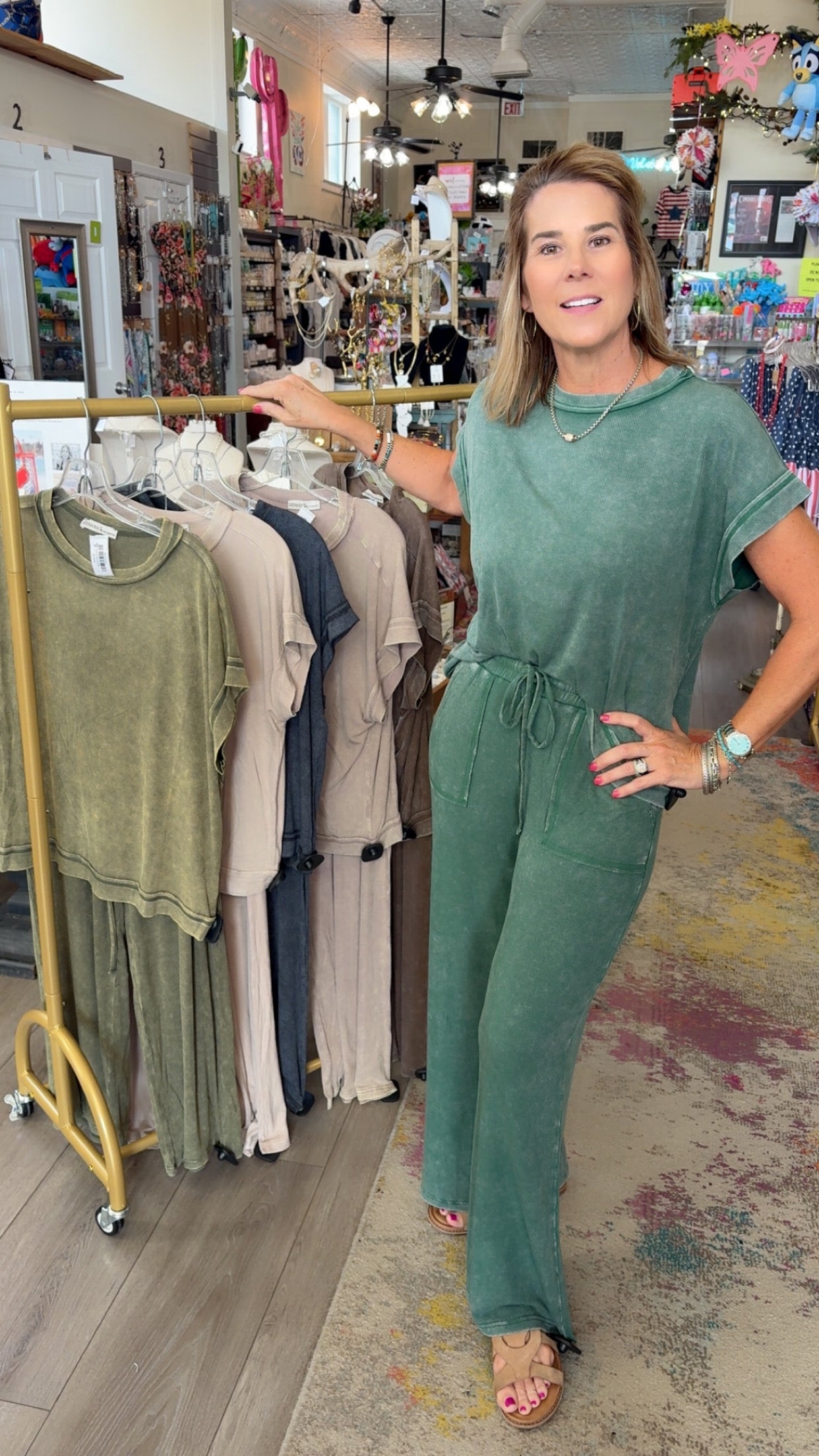 Woman in a green jumpsuit standing in a clothing store with racks of clothes in the background.