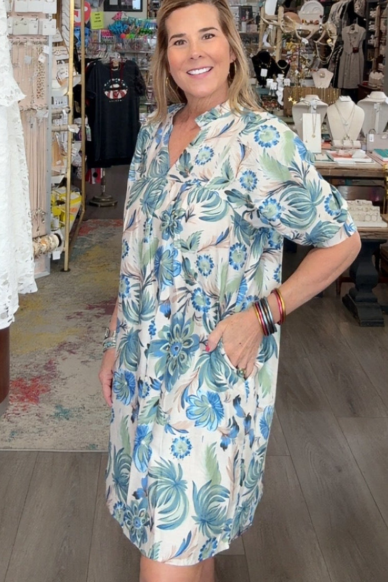Woman in a floral dress standing in a store with clothing and decor items around.
