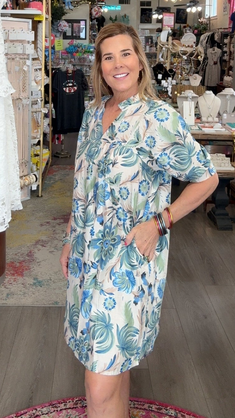 Woman in a floral dress standing in a store with clothing and decor items around.