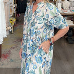 Woman in a floral dress standing in a store with clothing and decor items around.