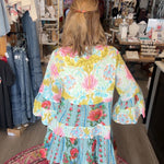 A woman viewed from behind wearing a colorful floral tiered dress inside a clothing boutique. The dress features a light blue base with pink, yellow, and green floral patterns and ruffled sleeves.