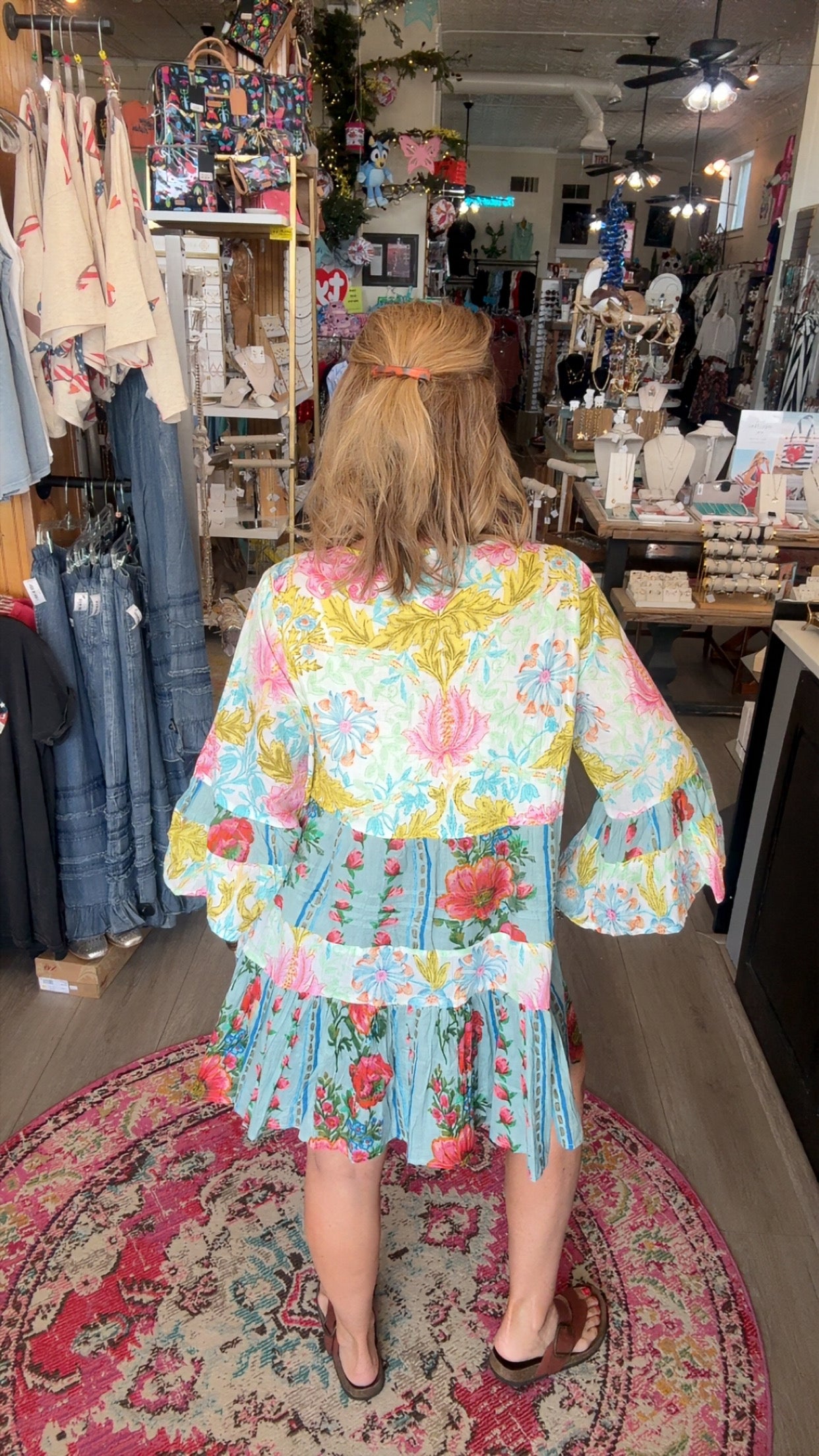 A woman viewed from behind wearing a colorful floral tiered dress inside a clothing boutique. The dress features a light blue base with pink, yellow, and green floral patterns and ruffled sleeves.