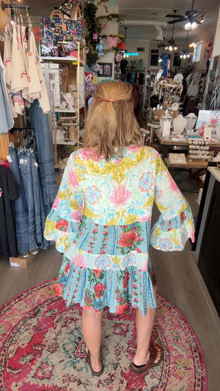 A woman viewed from behind wearing a colorful floral tiered dress inside a clothing boutique. The dress features a light blue base with pink, yellow, and green floral patterns and ruffled sleeves.