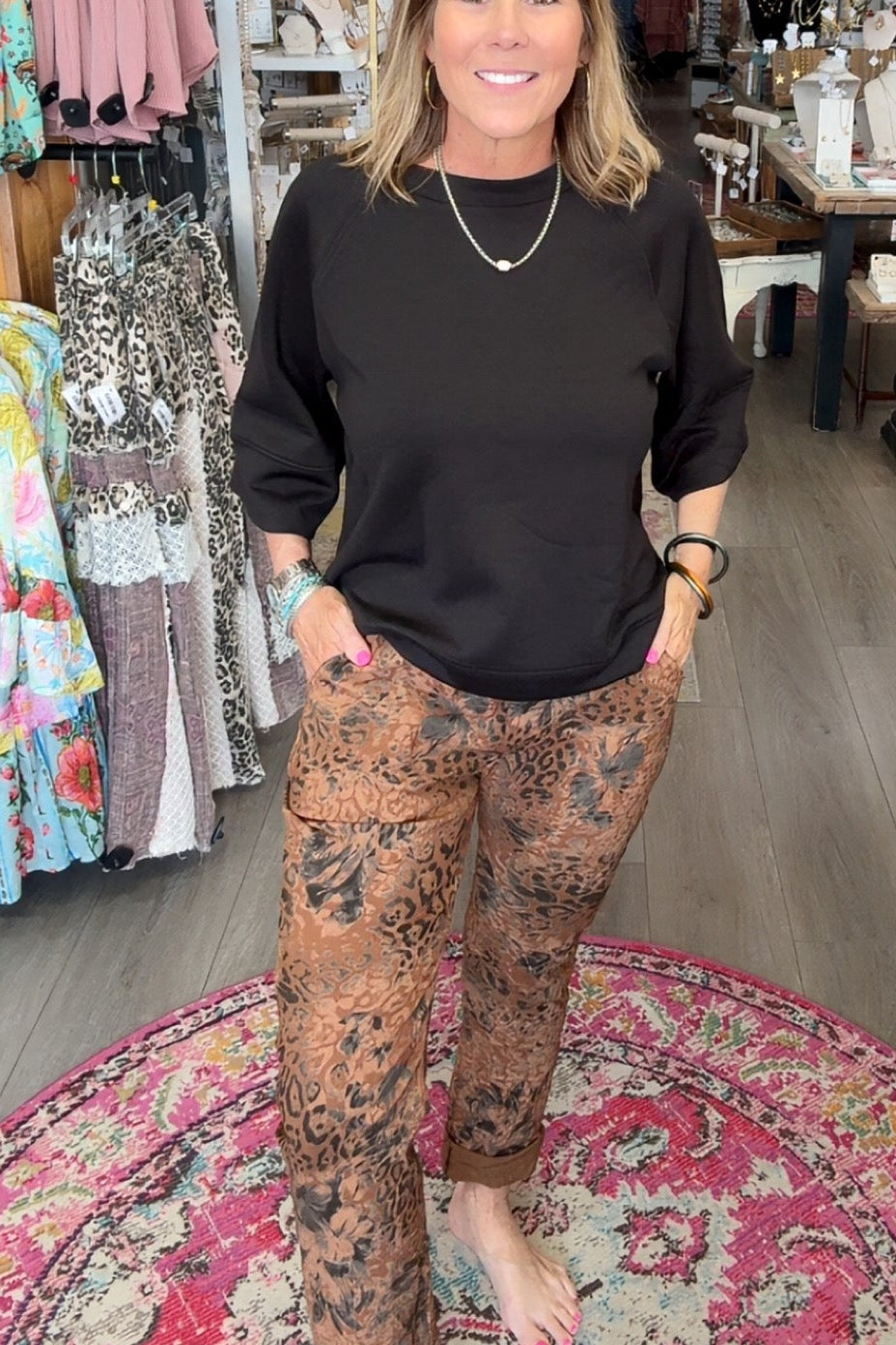 A woman stands in a boutique wearing a black long-sleeve top and brown patterned pants, standing barefoot on a round patterned rug.