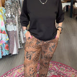 A woman stands in a boutique wearing a black long-sleeve top and brown patterned pants, standing barefoot on a round patterned rug.