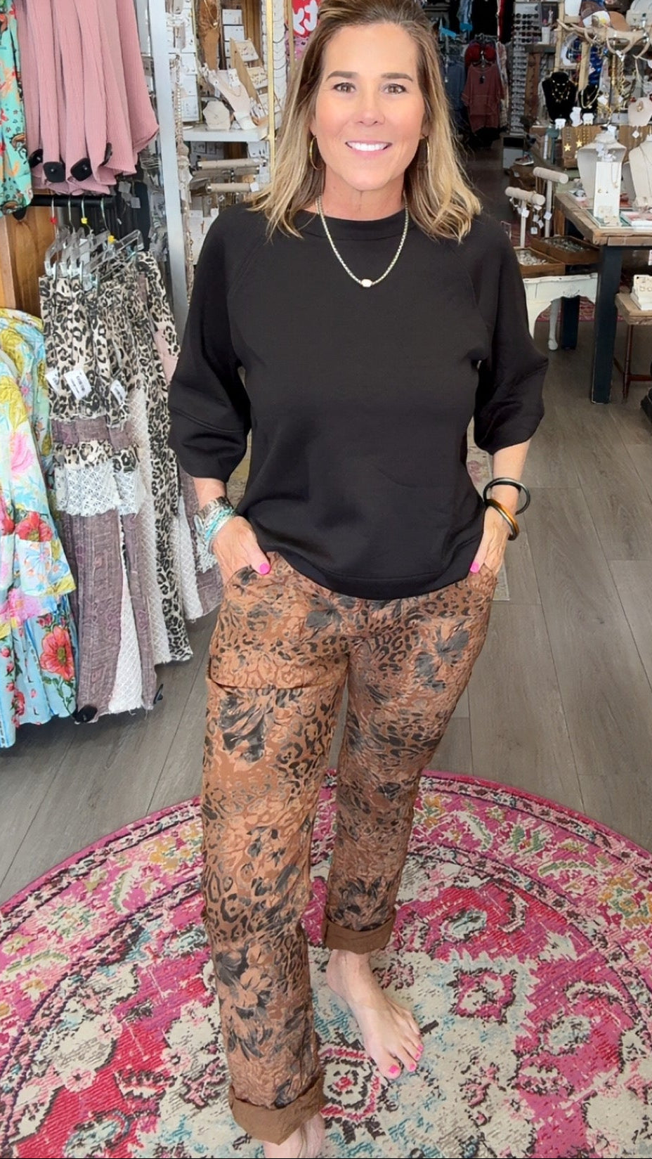 A woman stands in a boutique wearing a black long-sleeve top and brown patterned pants, standing barefoot on a round patterned rug.