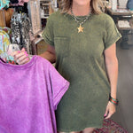 A woman standing in a boutique shop wearing an olive green short-sleeve t-shirt dress, holding up a purple tie-dye t-shirt on a hanger.