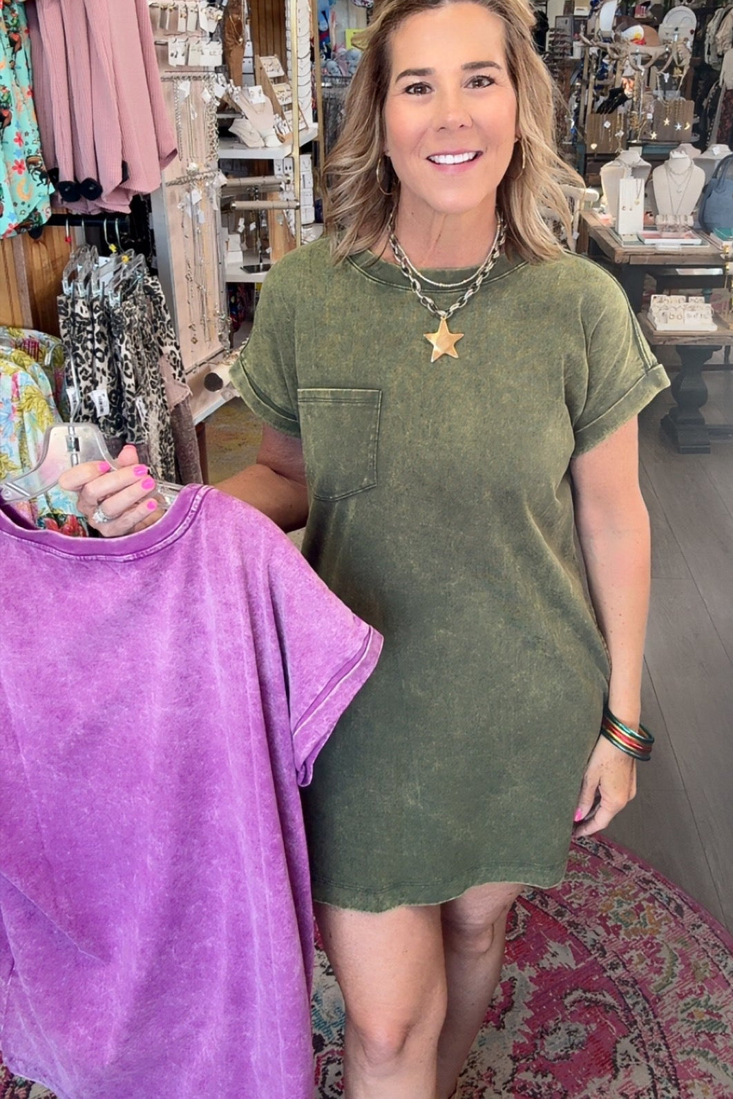 A woman standing in a boutique shop wearing an olive green short-sleeve t-shirt dress, holding up a purple tie-dye t-shirt on a hanger.