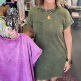 A woman standing in a boutique shop wearing an olive green short-sleeve t-shirt dress, holding up a purple tie-dye t-shirt on a hanger.