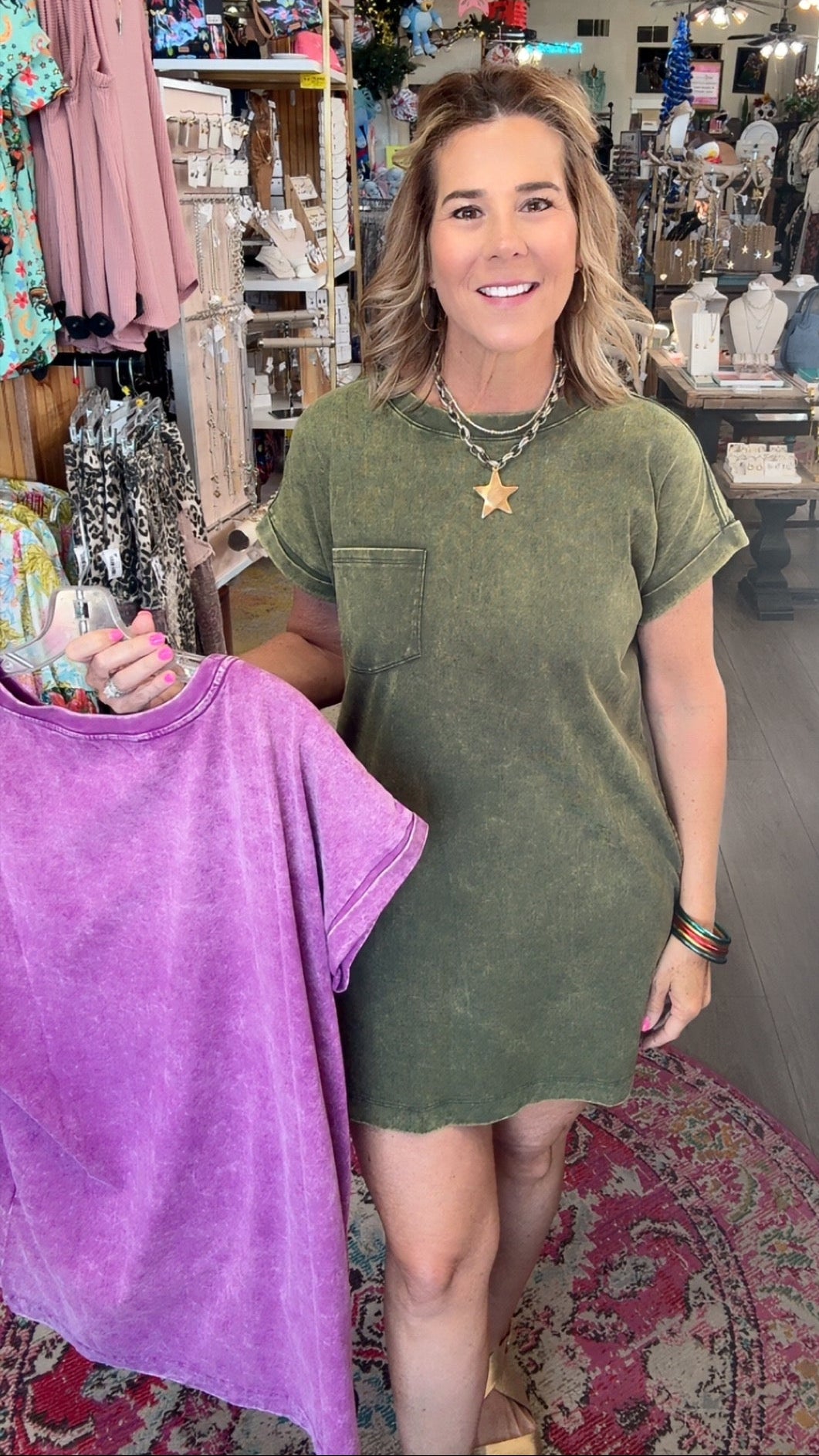 A woman standing in a boutique shop wearing an olive green short-sleeve t-shirt dress, holding up a purple tie-dye t-shirt on a hanger.