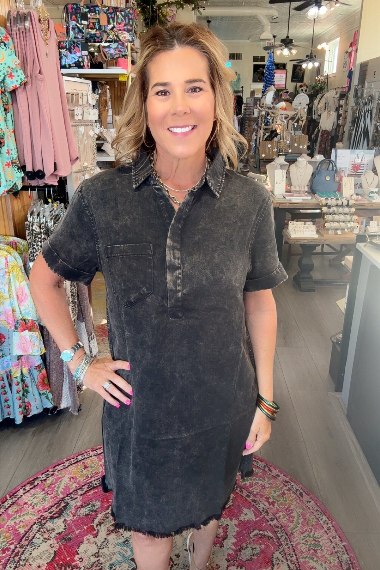 A woman modeling a short-sleeve, button-front shirt dress in a dark washed charcoal gray fabric, standing inside a boutique shop.