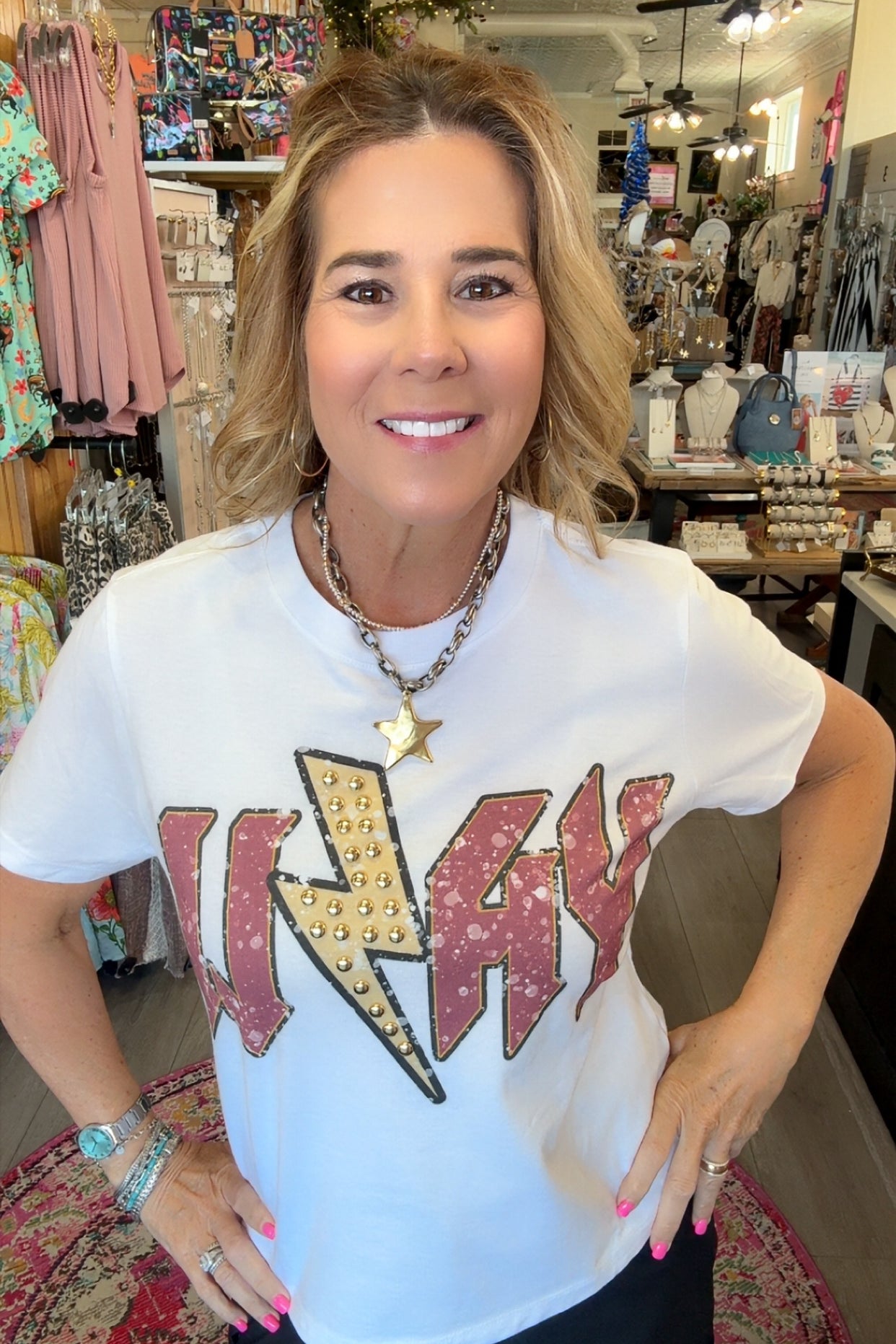 A woman wearing a white graphic t-shirt with a large red and gold 'WAYNE' text design featuring a lightning bolt, standing in a retail store.