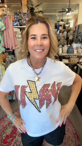 A woman wearing a white graphic t-shirt with a large red and gold 'WAYNE' text design featuring a lightning bolt, standing in a retail store.