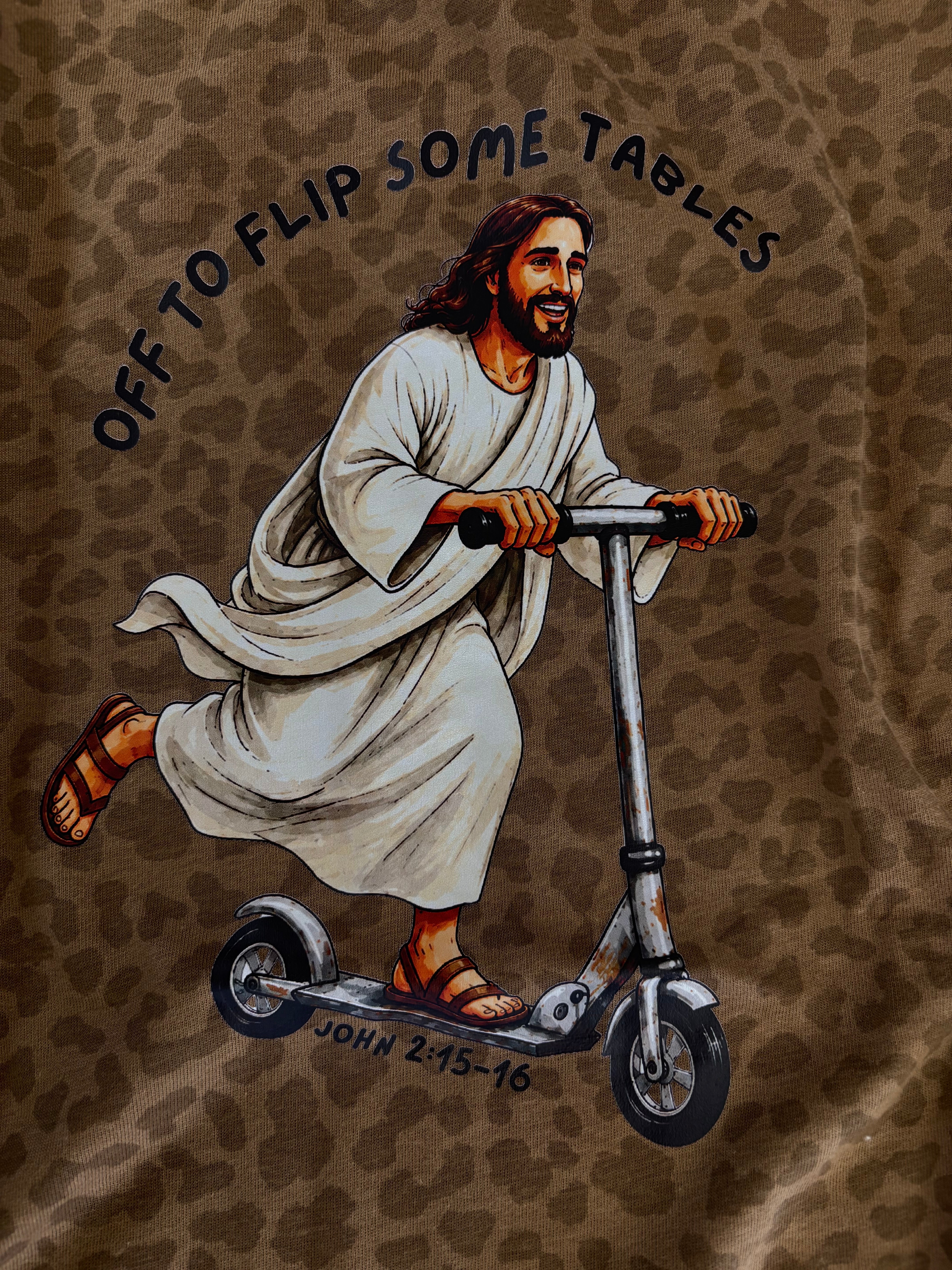 A close-up of a brown leopard-print fabric featuring a printed illustration of Jesus riding a scooter, with the text 'OFF TO FLIP SOME TABLES' and 'JOHN 2:15-16' printed on it.