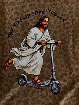 A close-up of a brown leopard-print fabric featuring a printed illustration of Jesus riding a scooter, with the text 'OFF TO FLIP SOME TABLES' and 'JOHN 2:15-16' printed on it.