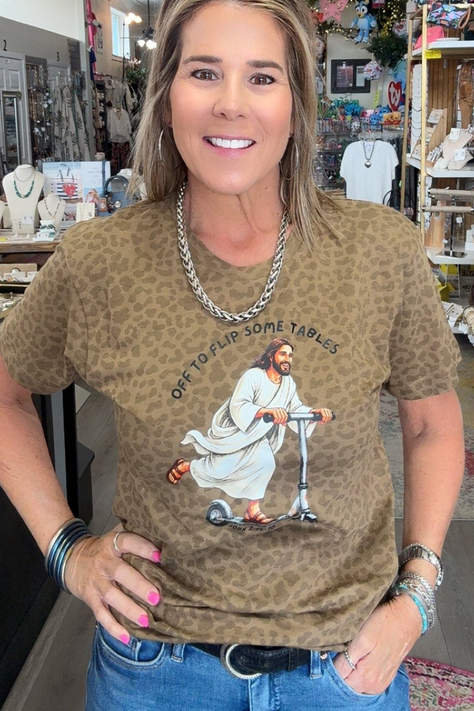 A woman wearing a brown leopard-print graphic t-shirt with a scooter illustration and the text 'EVERY BIRD WORTH A FEELING WRONG' paired with blue jeans, standing inside a retail store.