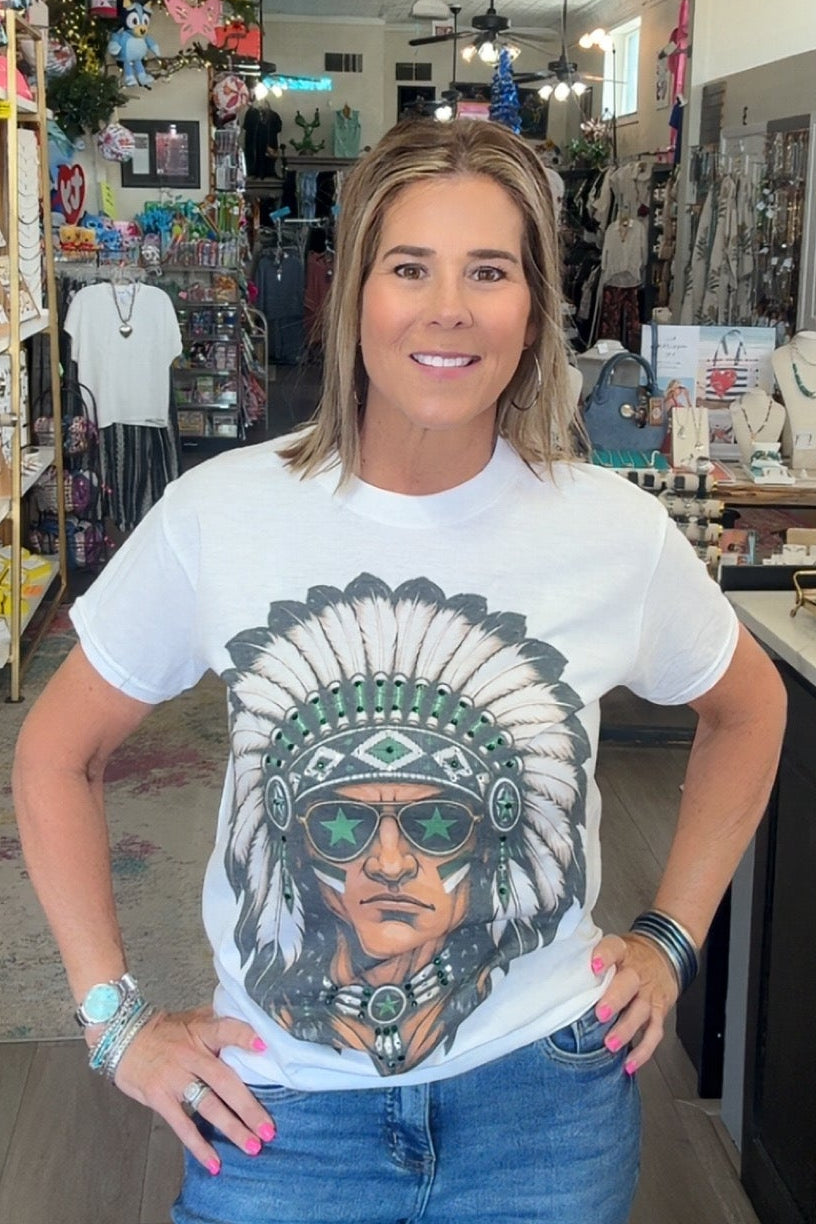 A woman stands inside a retail shop wearing a white graphic t-shirt with a detailed portrait of a Native American man in a feathered headdress and sunglasses, paired with blue jeans.