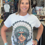 A woman stands inside a retail shop wearing a white graphic t-shirt with a detailed portrait of a Native American man in a feathered headdress and sunglasses, paired with blue jeans.