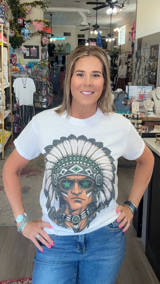 A woman stands inside a retail shop wearing a white graphic t-shirt with a detailed portrait of a Native American man in a feathered headdress and sunglasses, paired with blue jeans.