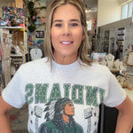 A woman wearing a light gray graphic t-shirt with 'INDIANYS' and a varsity jacket design in green and black, standing in a retail store.