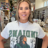 A woman wearing a light gray graphic t-shirt with 'INDIANYS' and a varsity jacket design in green and black, standing in a retail store.