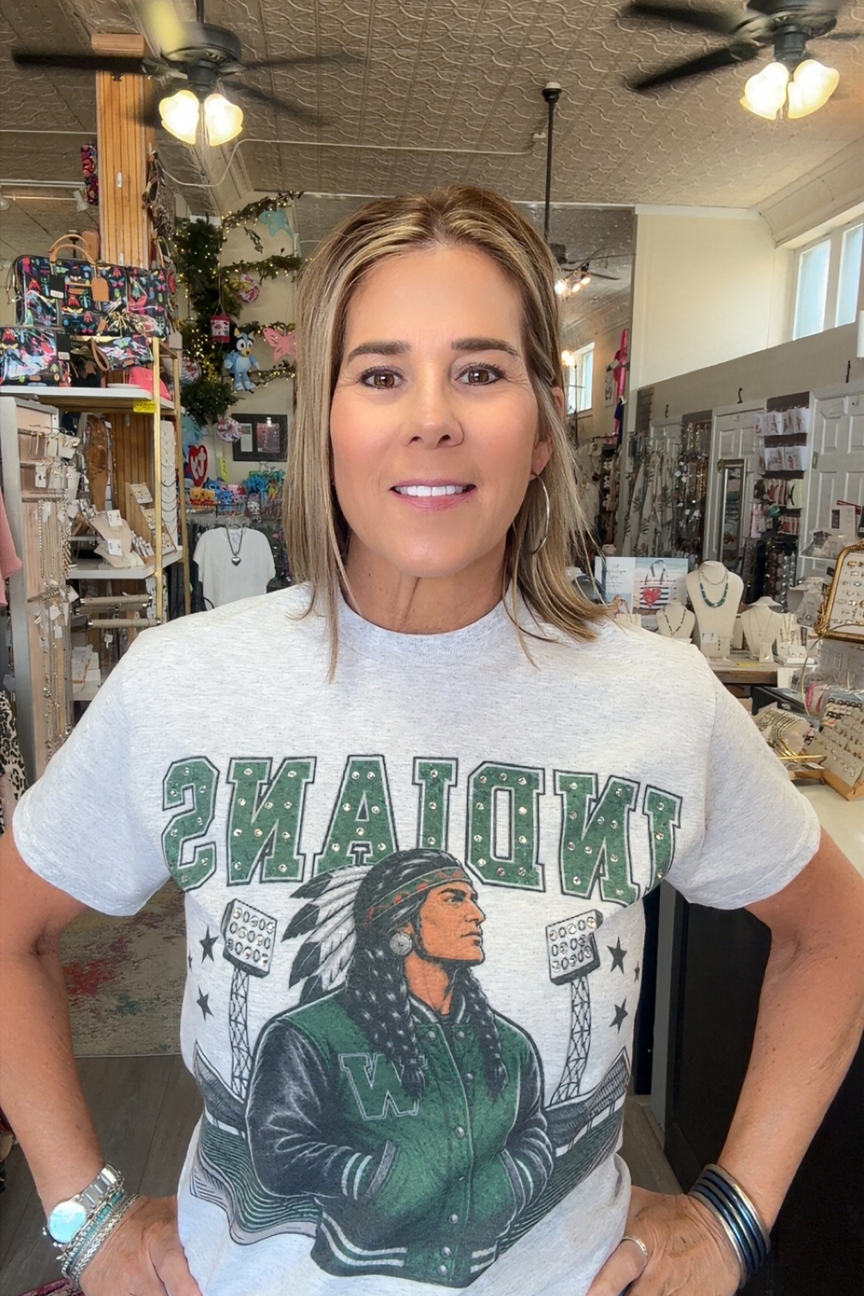 A woman wearing a light gray graphic t-shirt with 'INDIANYS' and a varsity jacket design in green and black, standing in a retail store.