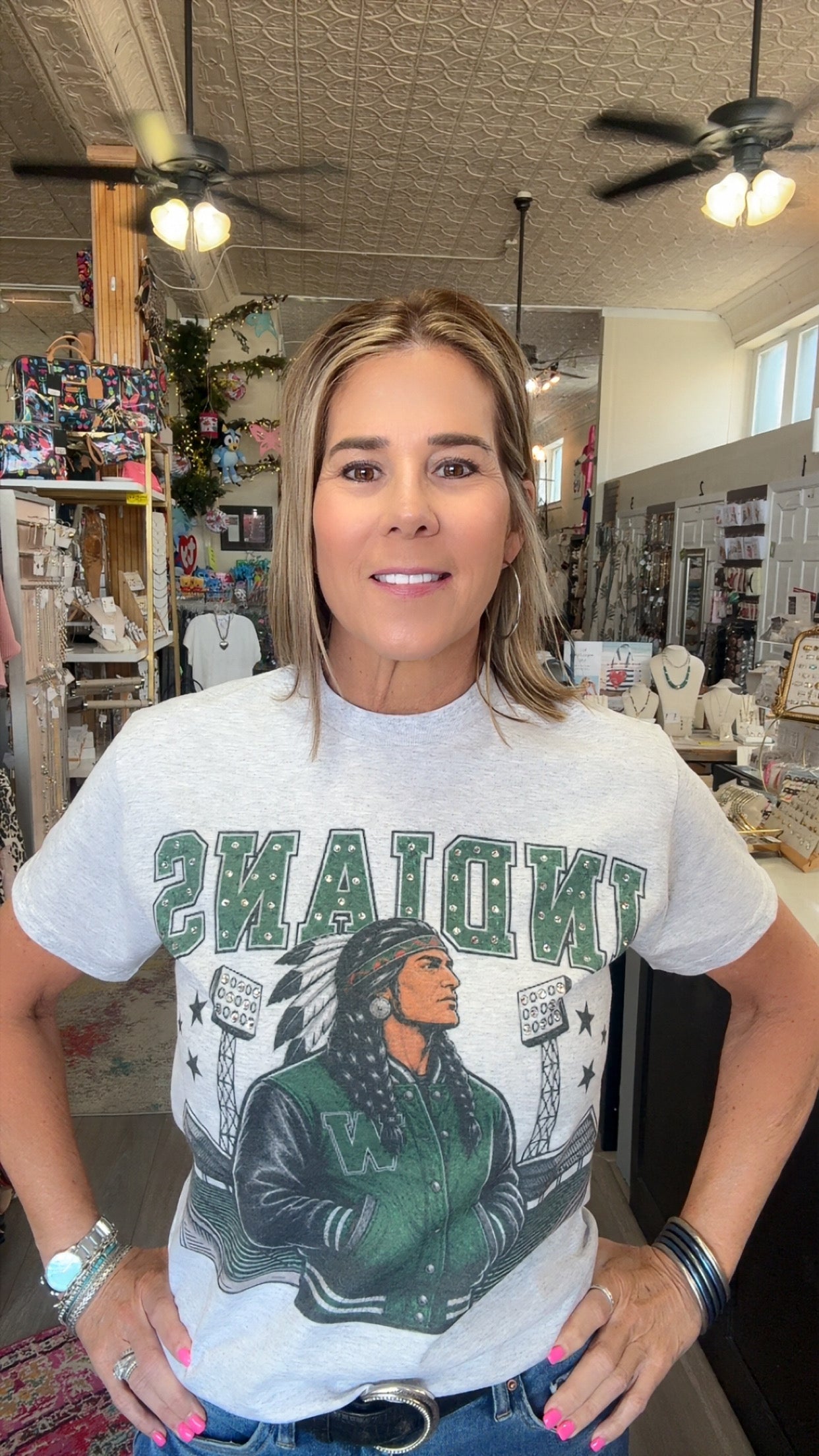 A woman wearing a light gray graphic t-shirt with 'INDIANYS' and a varsity jacket design in green and black, standing in a retail store.