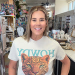 A woman wearing a white graphic t-shirt with a leopard print and turquoise lettering, standing inside a retail store.