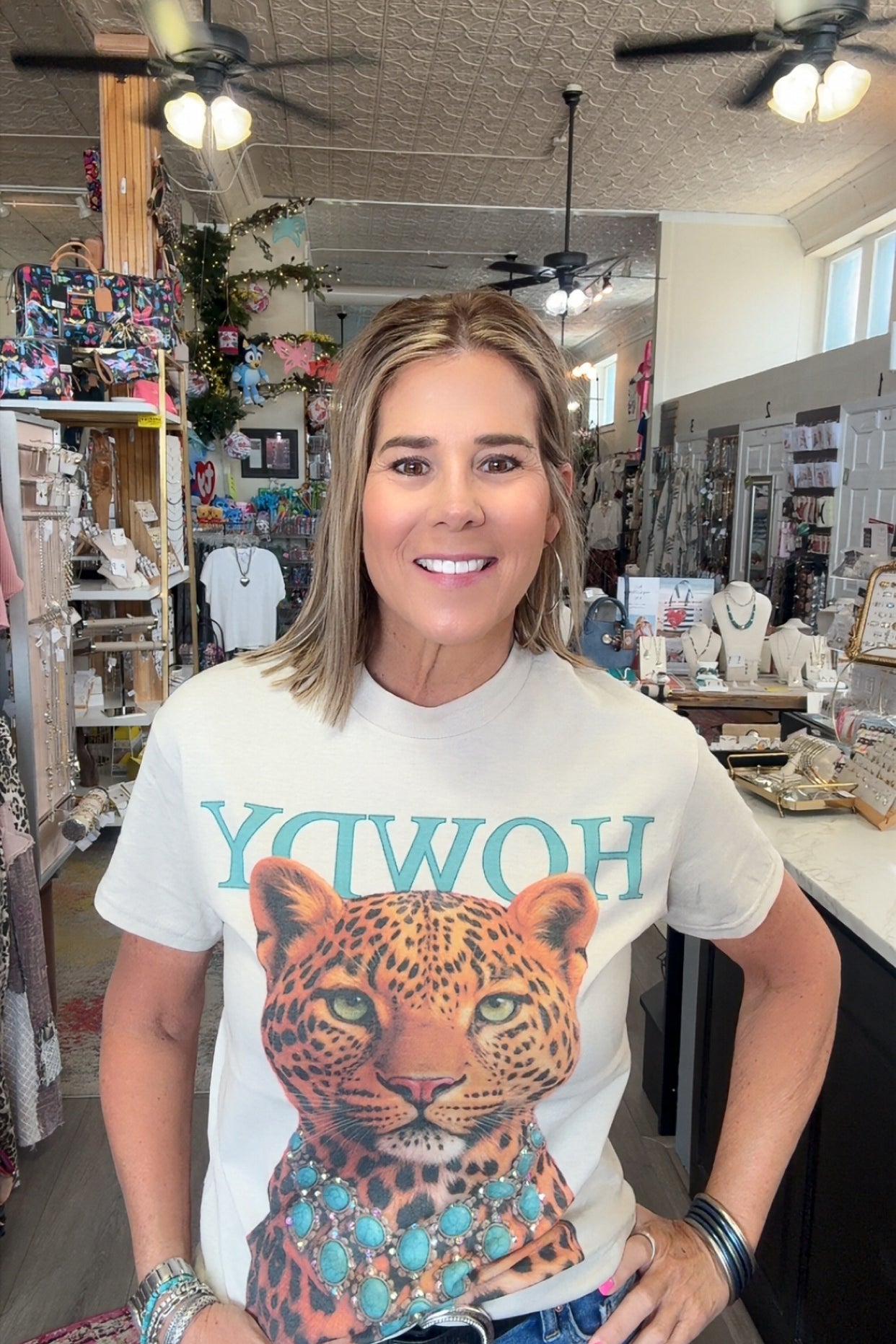 A woman wearing a white graphic t-shirt with a leopard print and turquoise lettering, standing inside a retail store.