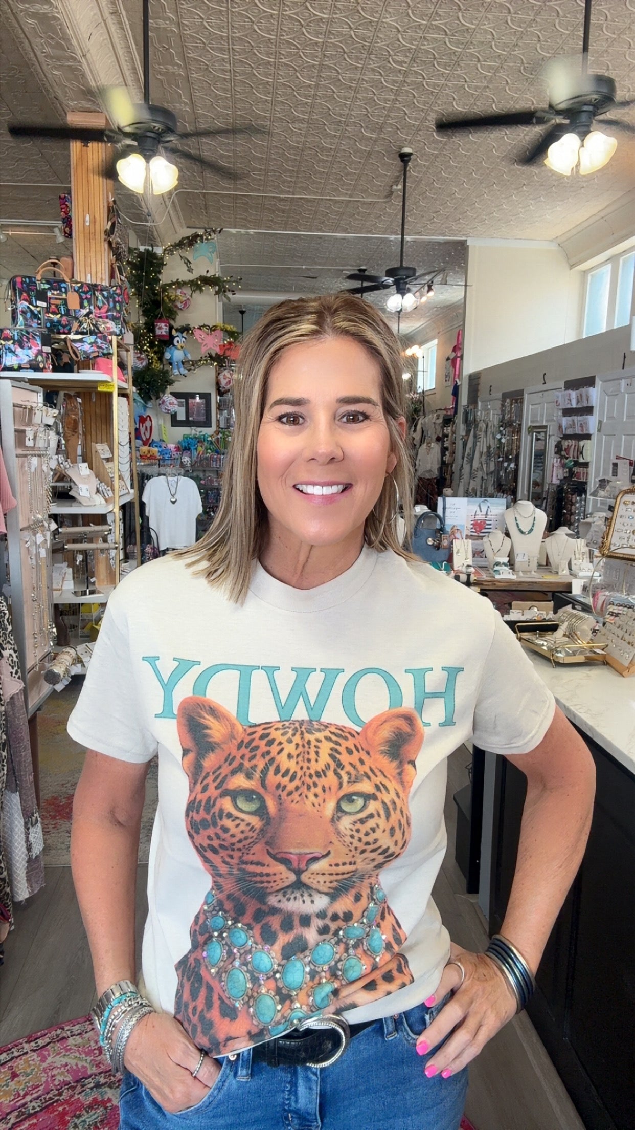 A woman wearing a white graphic t-shirt with a leopard print and turquoise lettering, standing inside a retail store.