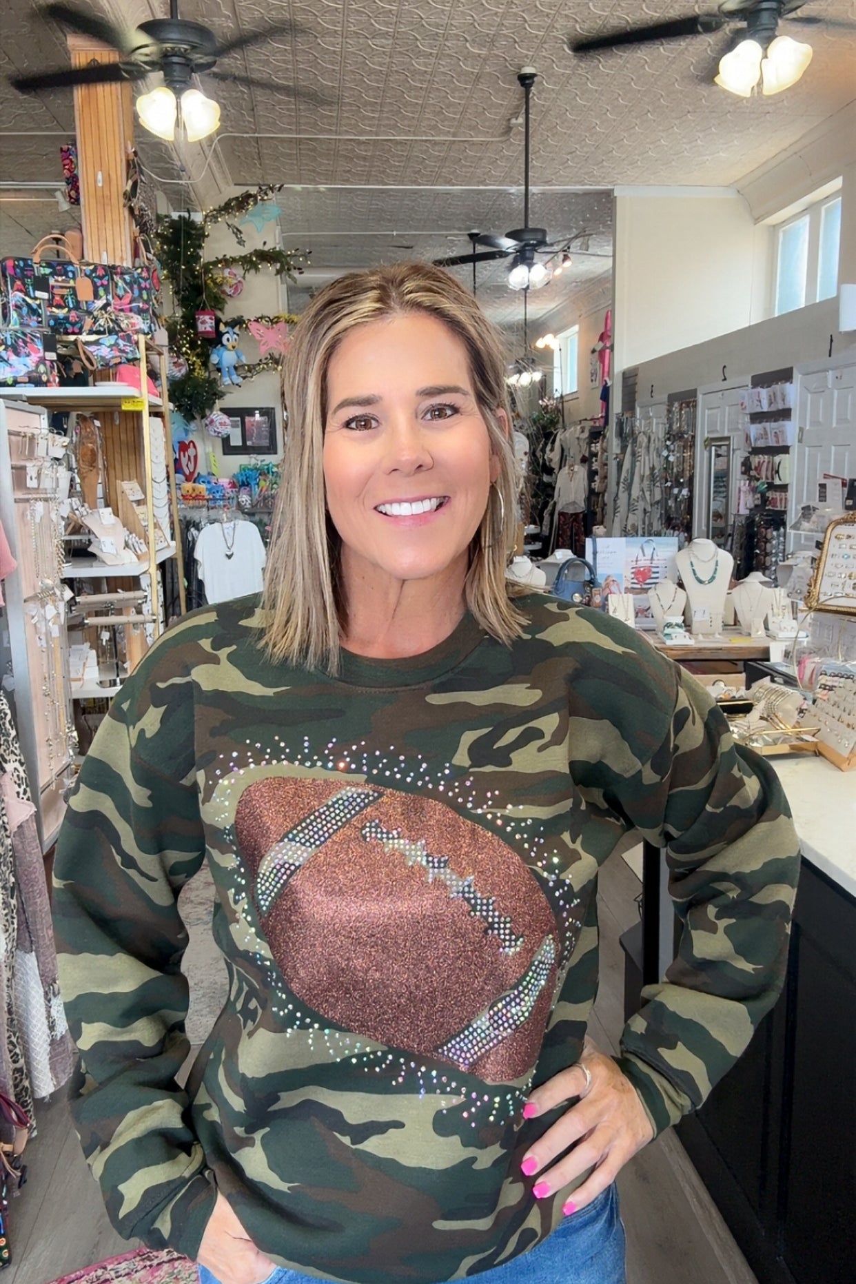 A woman wearing a camouflage crewneck sweatshirt with a large brown football graphic on the front, standing inside a retail shop.