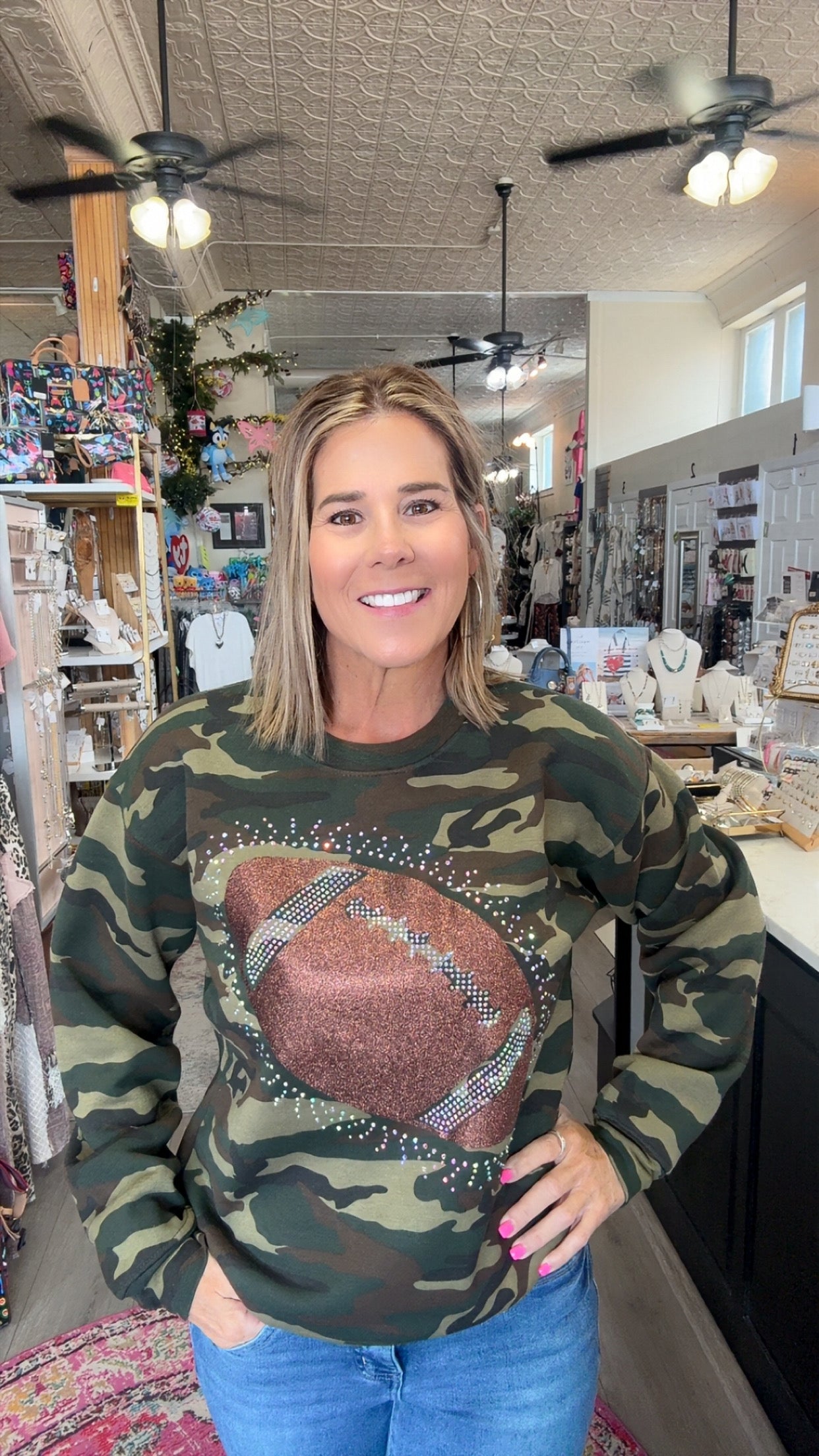 A woman wearing a camouflage crewneck sweatshirt with a large brown football graphic on the front, standing inside a retail shop.