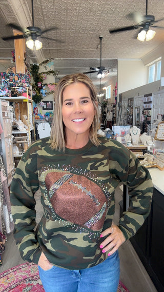 A woman wearing a camouflage crewneck sweatshirt with a large brown football graphic on the front, standing inside a retail shop.