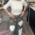 A woman posing in a boutique shop, wearing a cream-colored long-sleeve top and camouflage cargo pants with white heart-shaped patches.