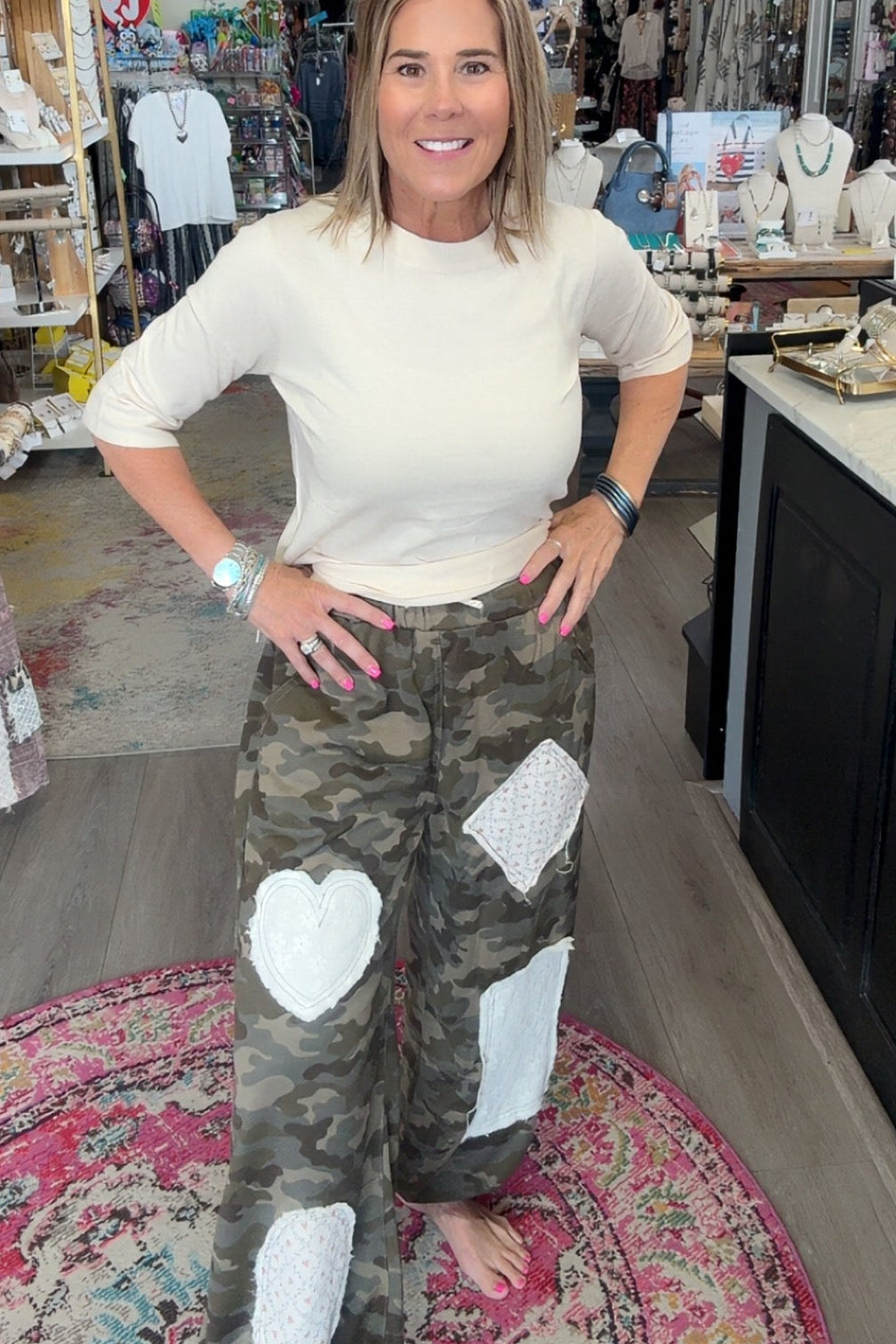 A woman posing in a boutique shop, wearing a cream-colored long-sleeve top and camouflage cargo pants with white heart-shaped patches.