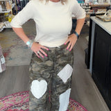 A woman posing in a boutique shop, wearing a cream-colored long-sleeve top and camouflage cargo pants with white heart-shaped patches.