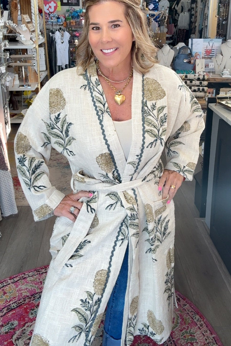 A woman stands inside a boutique shop wearing a cream-colored cotton robe with a green and gold botanical leaf pattern. The robe features a tie belt and is styled over a white top and blue jeans.