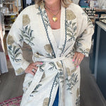 A woman stands inside a boutique shop wearing a cream-colored cotton robe with a green and gold botanical leaf pattern. The robe features a tie belt and is styled over a white top and blue jeans.