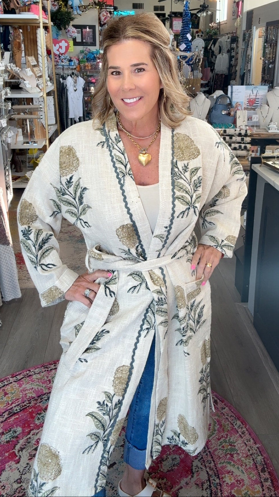 A woman stands inside a boutique shop wearing a cream-colored cotton robe with a green and gold botanical leaf pattern. The robe features a tie belt and is styled over a white top and blue jeans.
