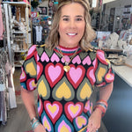 A woman wearing a colorful short-sleeve knit top with a heart pattern, standing in a retail store.