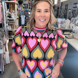 A woman wearing a colorful short-sleeve knit top with a heart pattern, standing in a retail store.